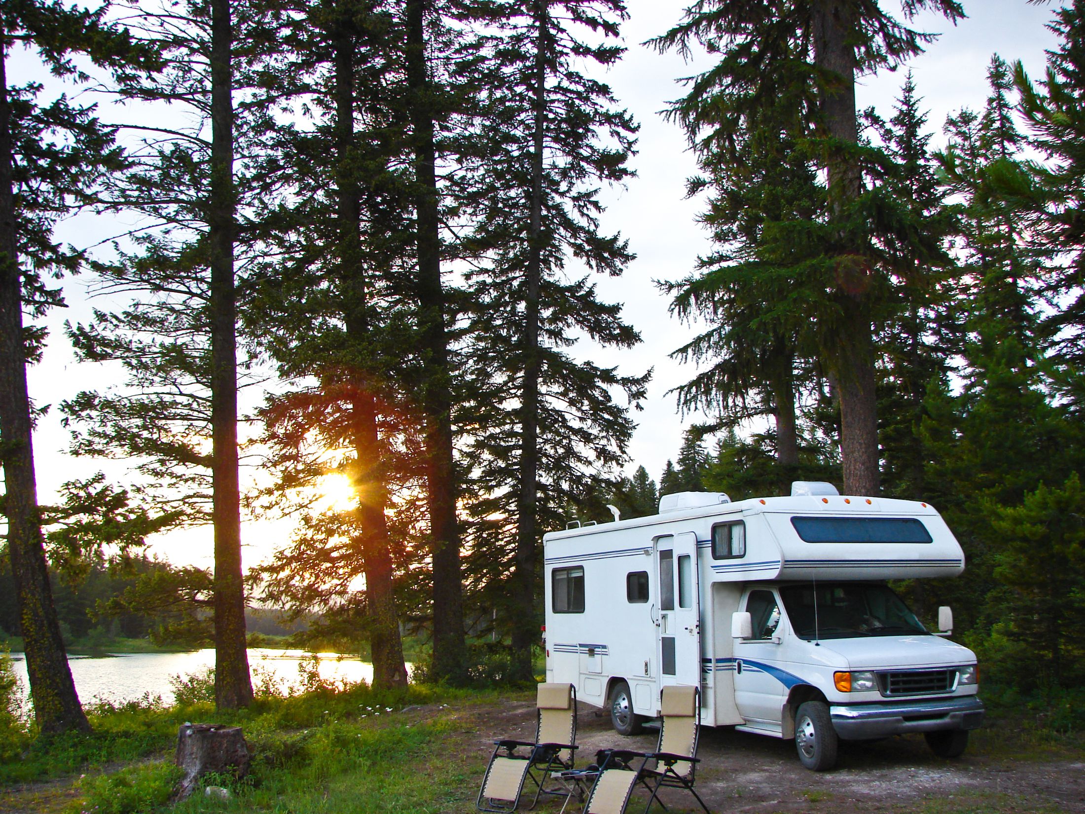 White motorhome parked by lake at sunset surrounded by tall evergreen trees with camping chairs in foreground